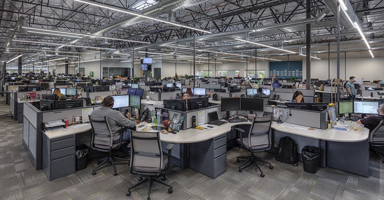 Numerous office employees are seated in cubicles in a busy office space.