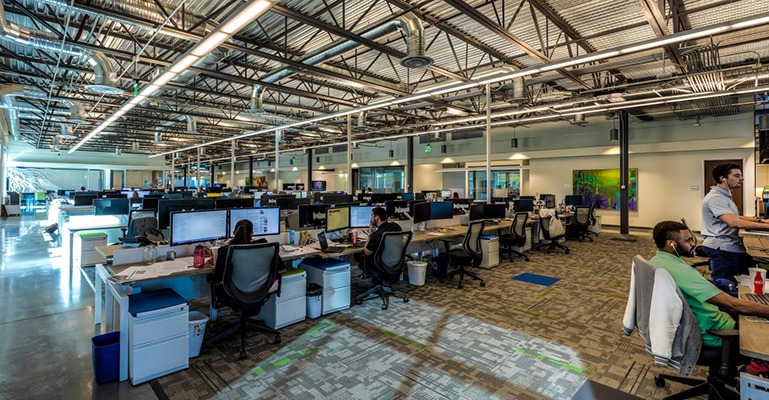 Numerous office employees are seated in cubicles with several others at shared desks off to one side.