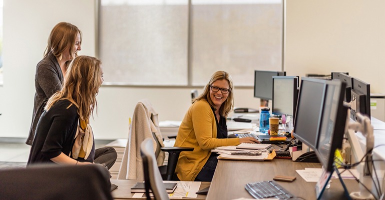 A female office worker seated at her desk looks over her shoulder to smile and laugh with two colleagues behind her.