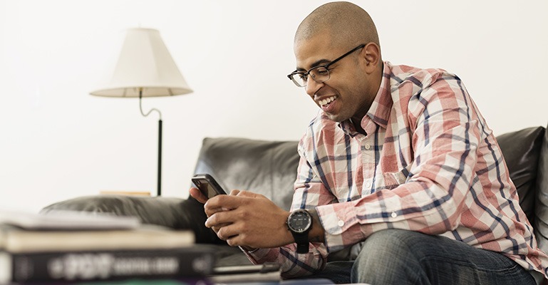 A smiling man sits on a couch, looking at his smartphone screen.