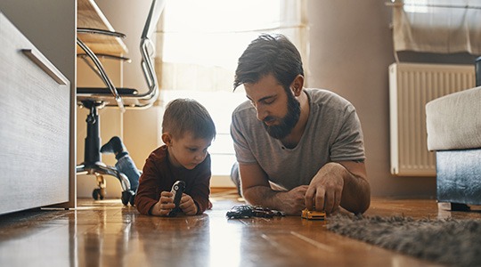 A young father and his small child play with toy cars on the floor.