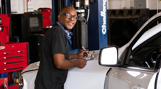 A bespectacled mechanic writes on a clipboard placed on the hood of a white car in a garage.