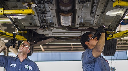 Two mechanics with flashlights work on the underside of a large vehicle.
