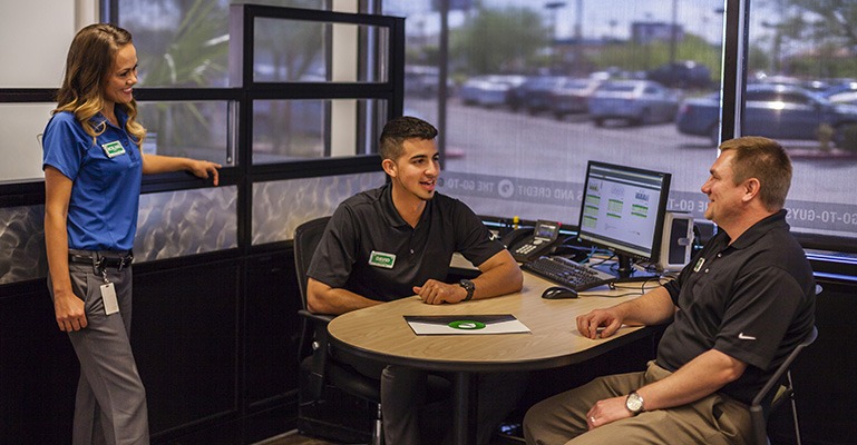 Three smiling customer service representatives are gathered around a desk, having a conversation between helping customers.